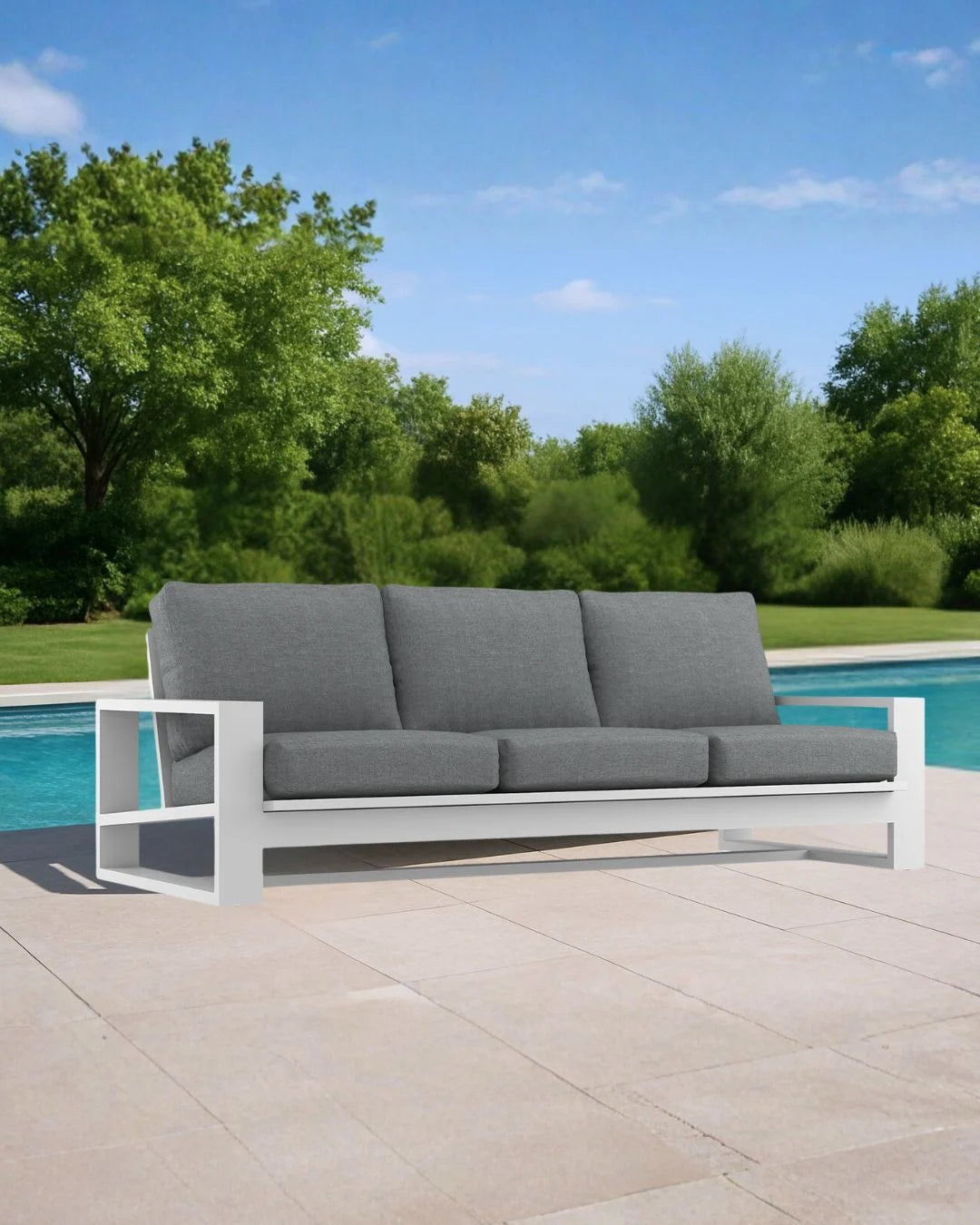 Gray outdoor sofa with white frame on a patio near a pool with greenery in the background