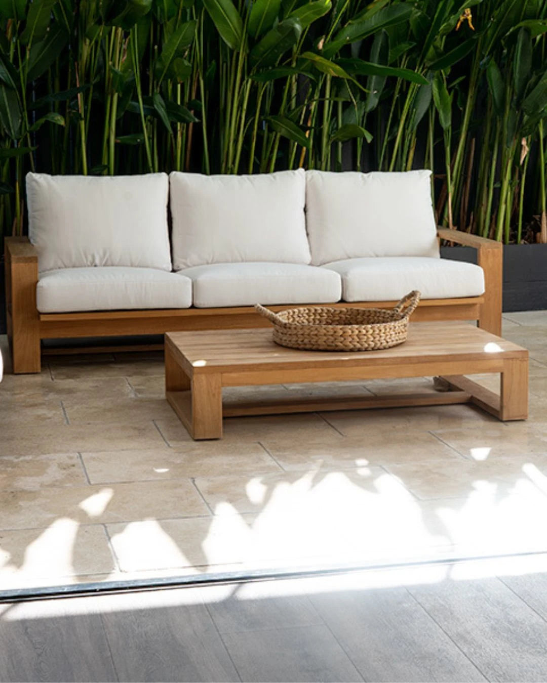 Wooden outdoor sofa with white cushions and a woven coffee table in front, set against a green foliage background.