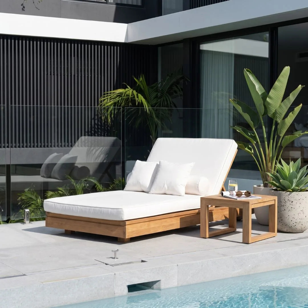 A modern outdoor chaise lounge with a white cushion and wooden frame sits beside a pool on a patio, styled with a matching wooden side table, potted plants, and a contemporary building in the background.