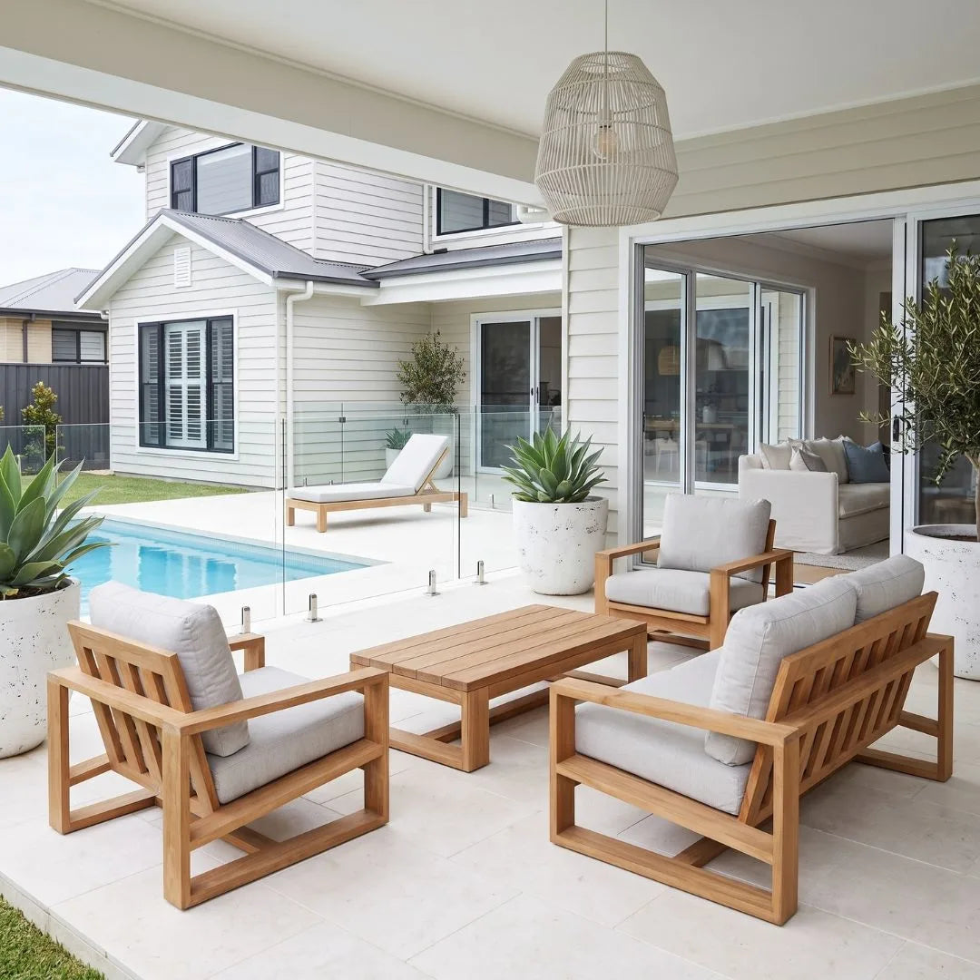 A modern outdoor patio furniture set featuring light natural wood frames and light gray cushions, arranged on a tiled patio under a covered porch with a swimming pool and house in the background.