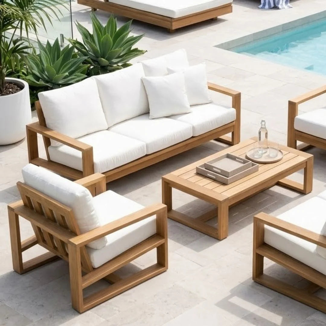 Outdoor patio furniture set with a wooden three-seat sofa, two matching armchairs, and a slatted coffee table, all with thick white cushions, arranged on a light stone deck near a pool.