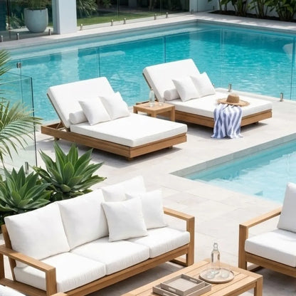 Outdoor poolside patio furniture set with white cushions and light wood frames, including a chaise lounge, armchair, and sofa, arranged beside a turquoise swimming pool with glass railings and plants.