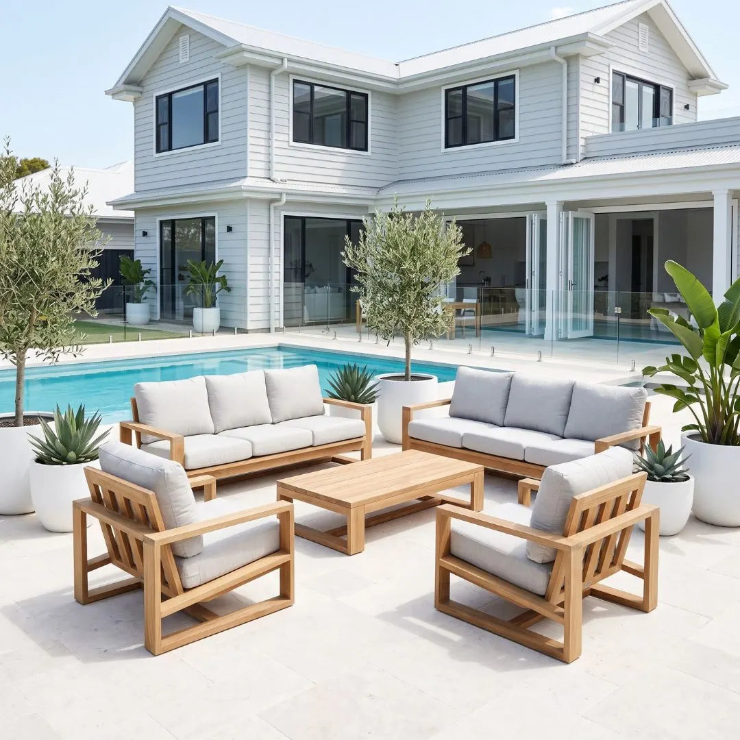 A 5-piece outdoor patio furniture set with light oak wood frames and light gray cushions, arranged on a tiled patio in front of a modern white house with a swimming pool.
