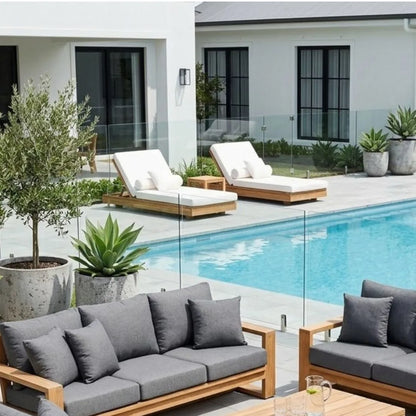 Modern outdoor patio furniture set with gray cushions and light wood frames, arranged beside a swimming pool in front of a white house with glass railings.
