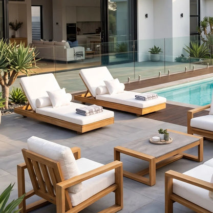 Outdoor patio furniture set with light wood frames and white cushions, including two chaise lounges, two armchairs, and a low rectangular coffee table, arranged beside a swimming pool in a modern villa setting.