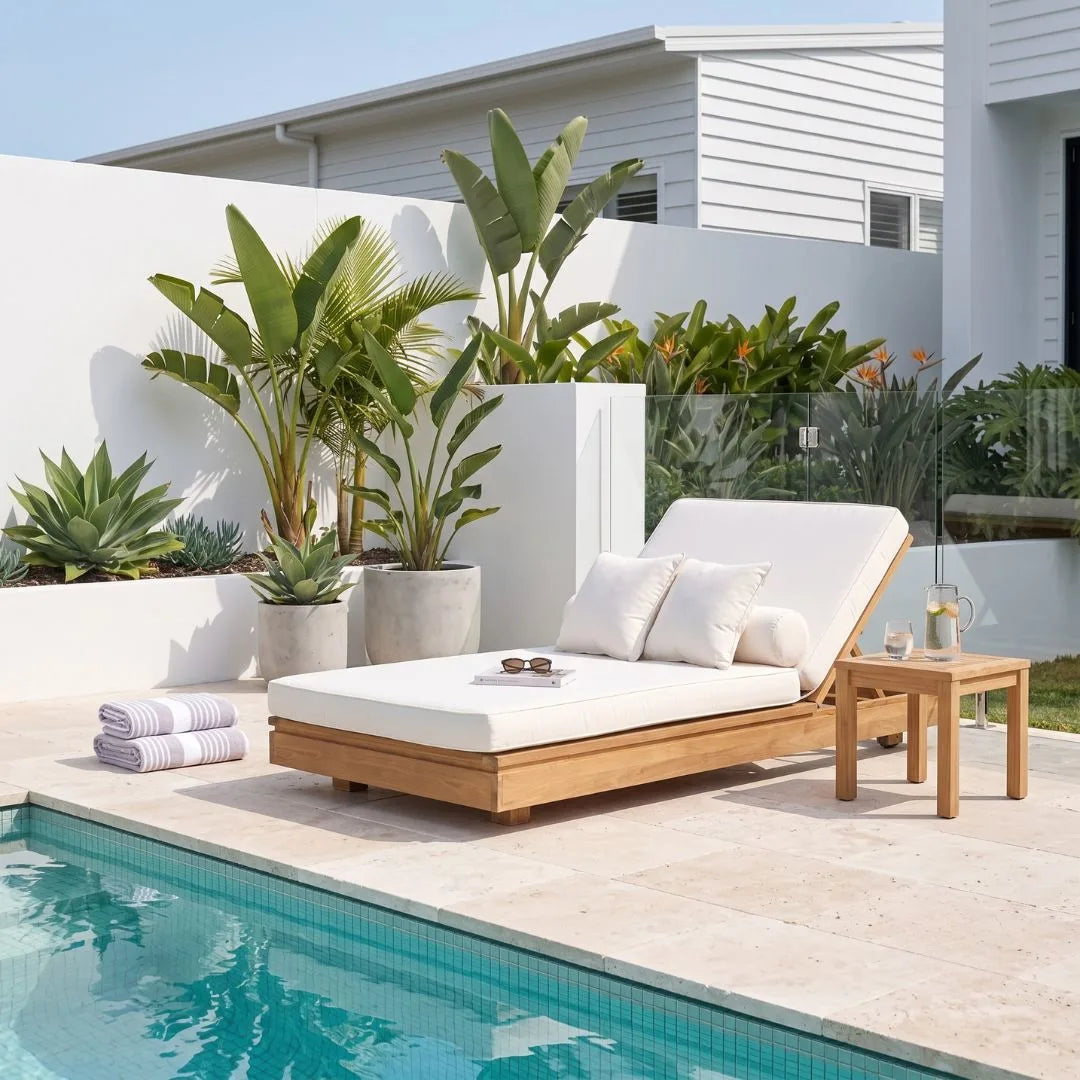 A modern outdoor chaise lounge with a light wood frame and white cushion sits beside a swimming pool on a tiled patio. The lounge is styled with white pillows, a small wooden side table, folded striped towels, and large potted tropical plants against a white wall and house background.