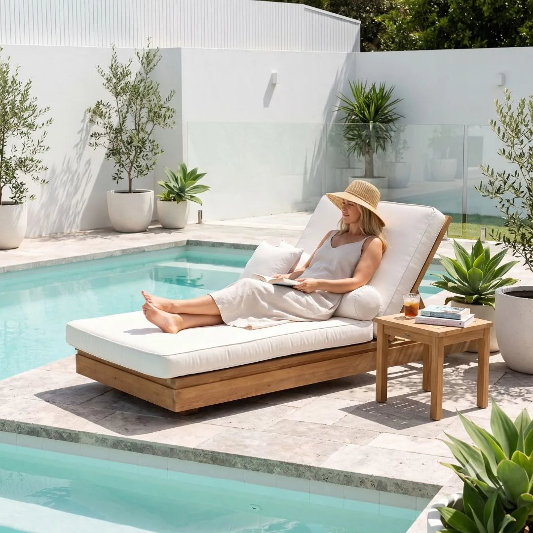 A woman relaxes on a wooden outdoor chaise lounge beside a swimming pool, wearing a straw hat and light dress. A small wooden side table sits next to the lounge, and potted plants and a modern white wall are in the background.