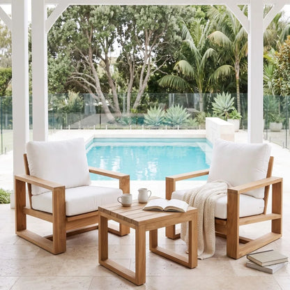 Two wooden outdoor armchairs with white cushions and a matching wooden side table, arranged on a patio overlooking a swimming pool and tropical greenery.