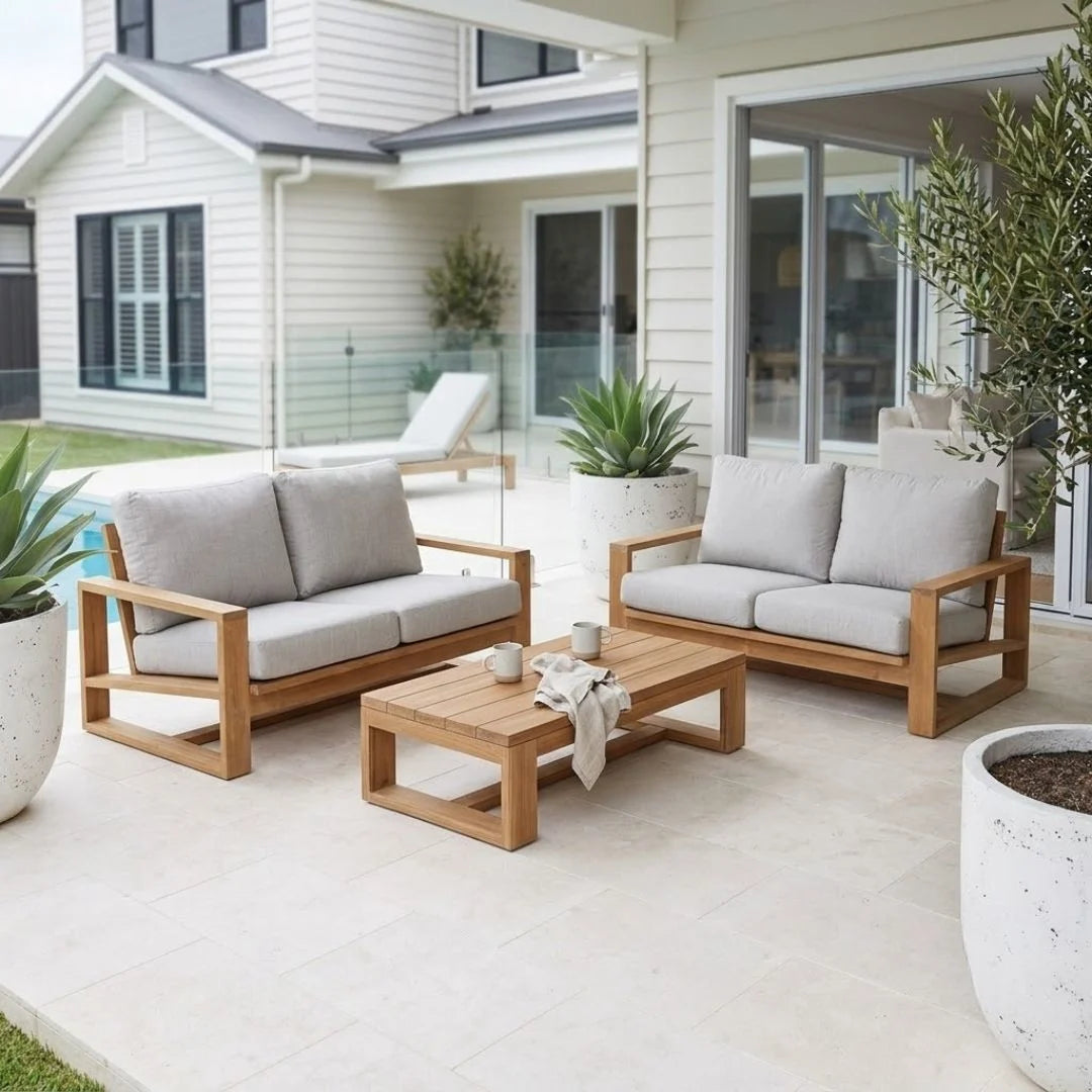 Outdoor patio furniture set including a two-seater sofa, a loveseat, and a rectangular coffee table in light natural wood with off-white cushions, arranged on a tiled patio in front of a modern house.