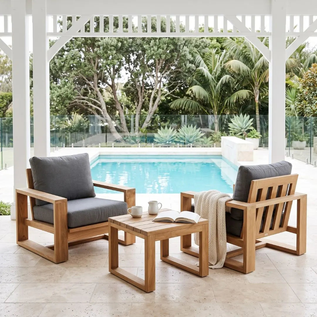 Two wooden outdoor patio armchairs with dark gray cushions and a matching wooden coffee table, arranged on a tiled patio under a white pergola overlooking a swimming pool and tropical garden.