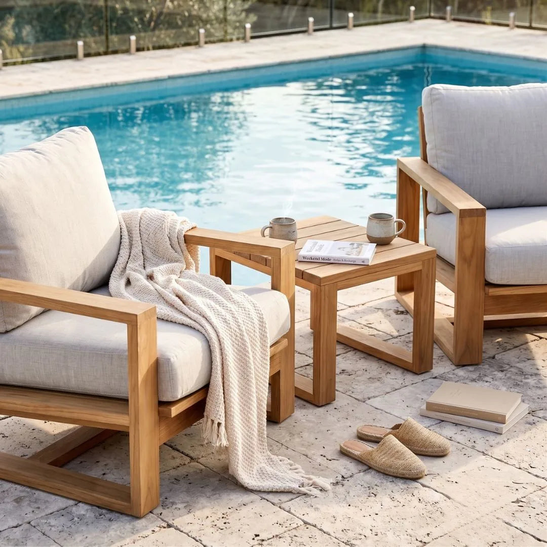 Outdoor patio furniture set with two light wood armchairs, a matching square side table, a cream throw blanket, mugs, a newspaper, and slippers beside a swimming pool.