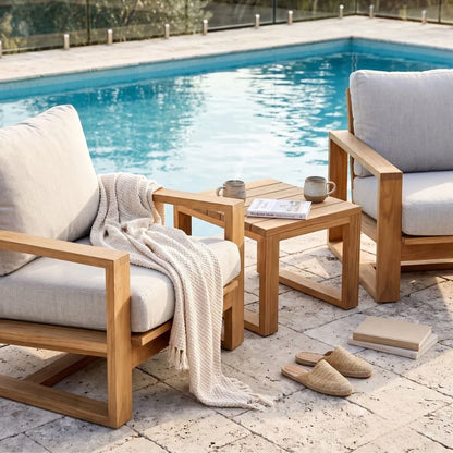 Outdoor patio furniture set with two light wood armchairs, a matching square side table, a cream throw blanket, mugs, a newspaper, and slippers beside a swimming pool.