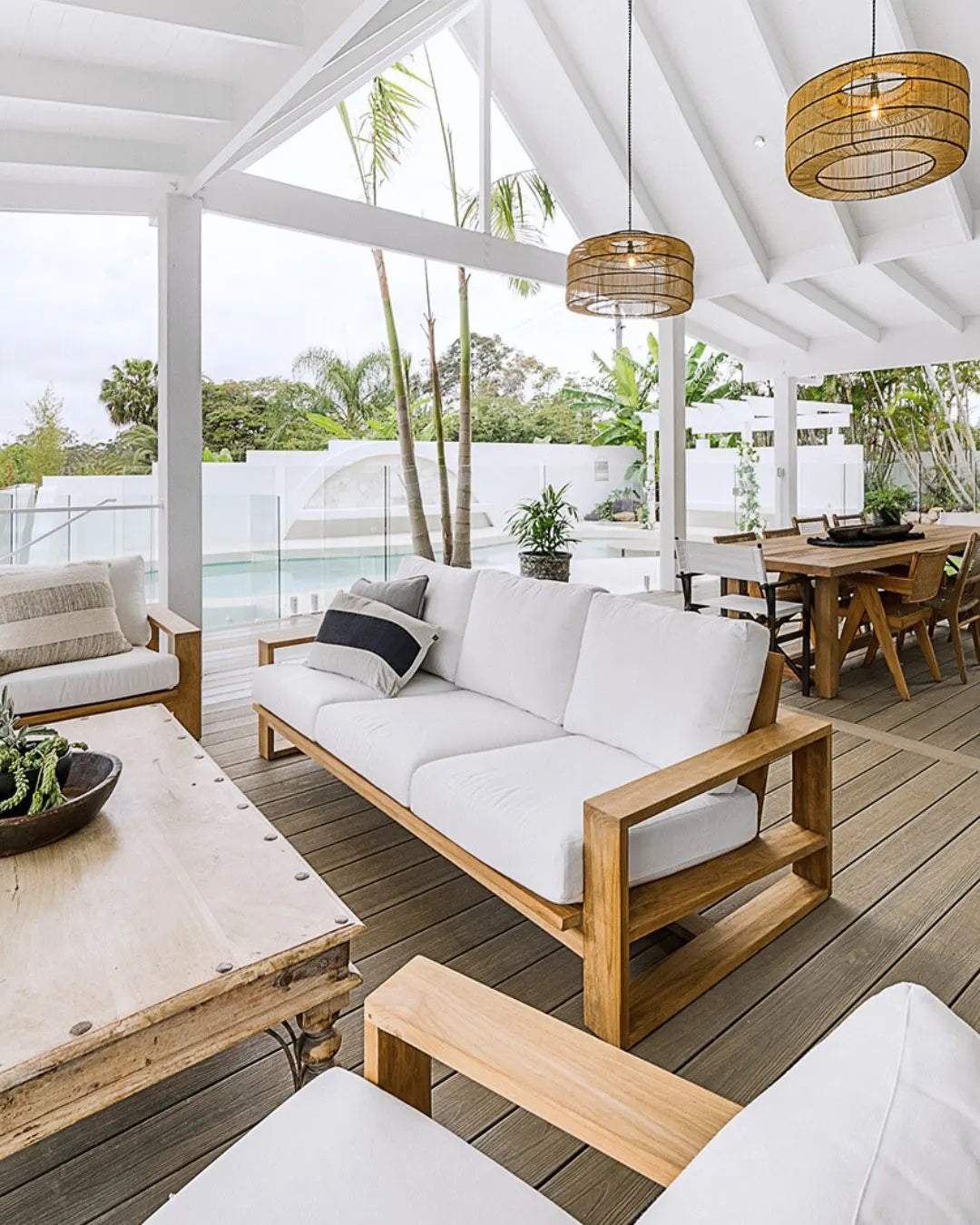 Outdoor patio with wooden furniture, white cushions, and a view of trees and a pool.