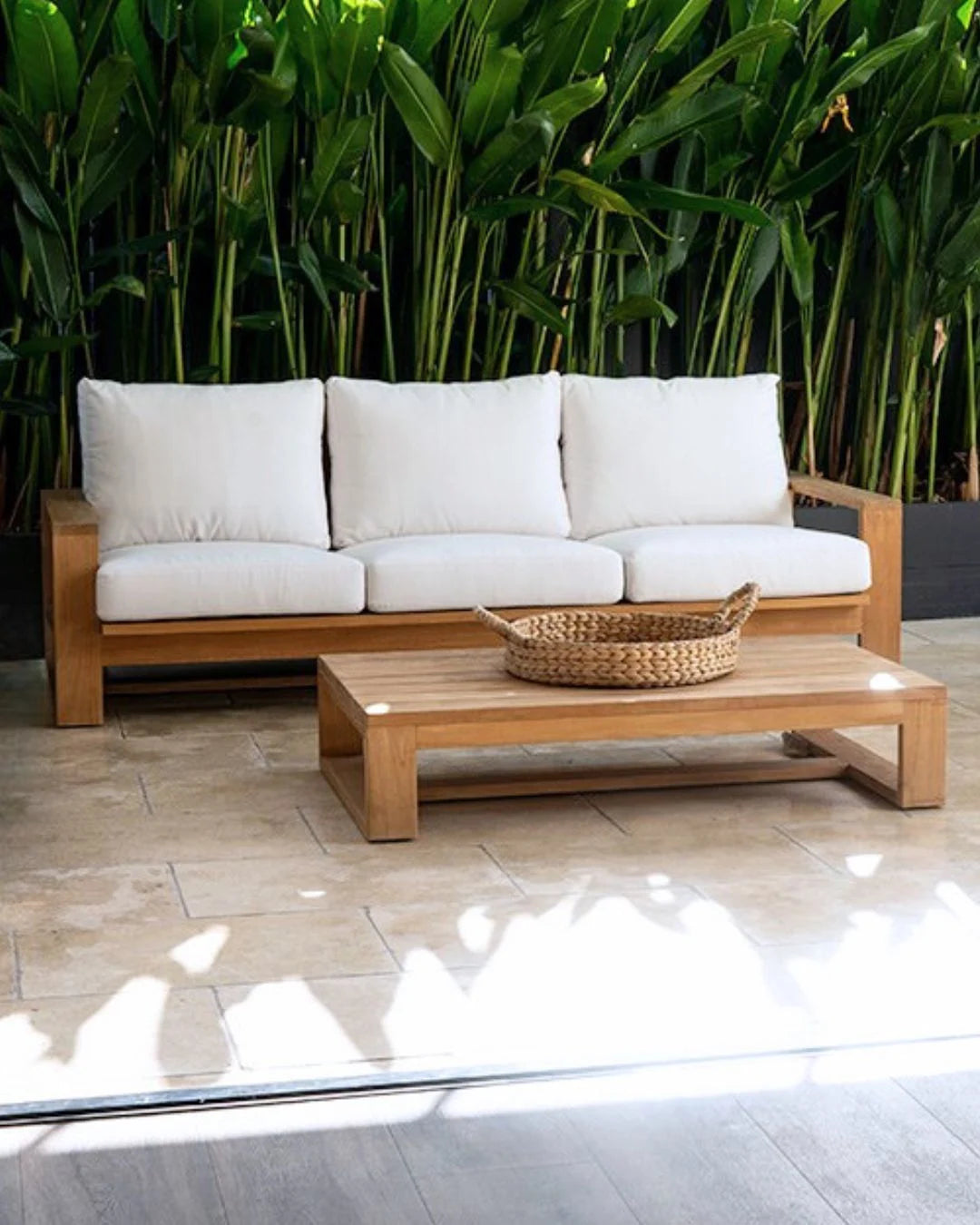 Wooden outdoor sofa with white cushions and a woven basket on a small table, set against a green foliage background.