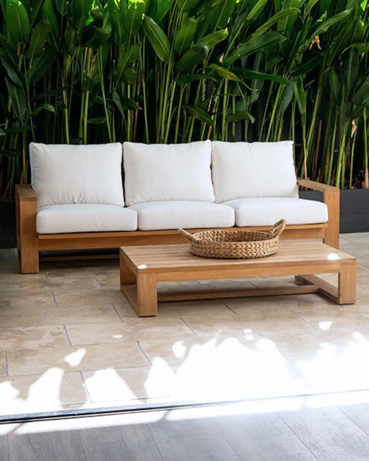 Wooden outdoor sofa with white cushions and a woven basket on a small table, set against a green foliage background.