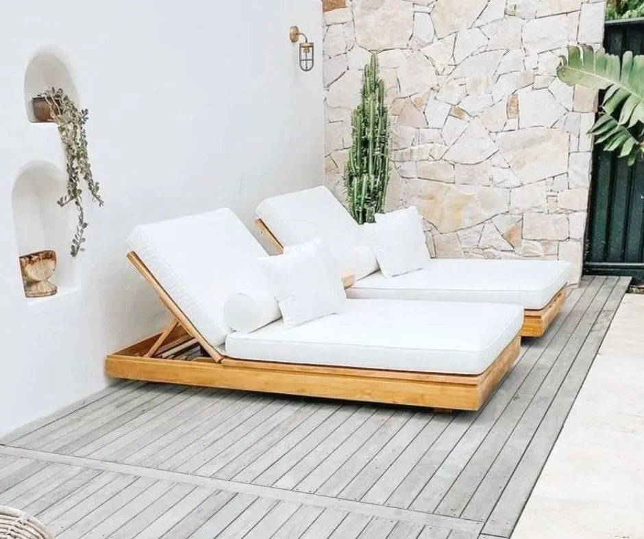 Outdoor sun lounge daybeds with white cushions on a wooden deck against a stone wall.