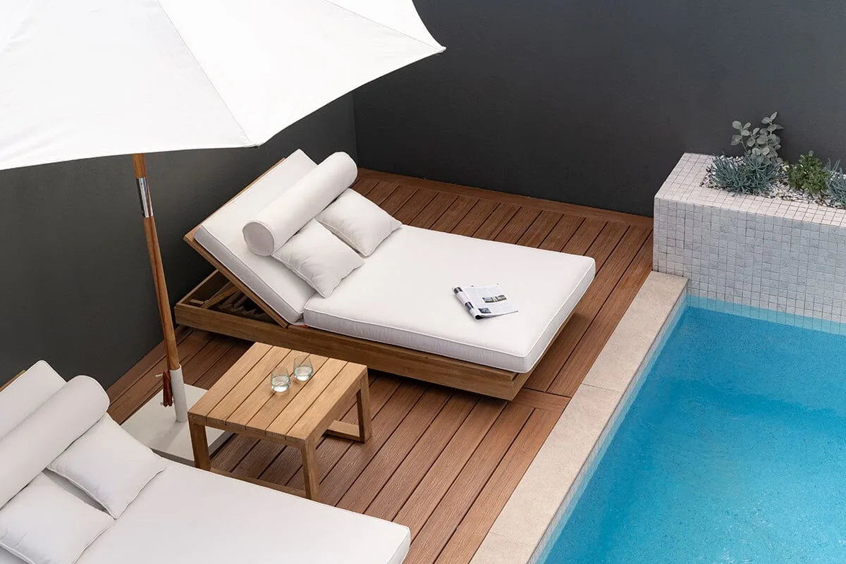 Outdoor sunlounge daybed with white cushions on a wooden deck by a pool.