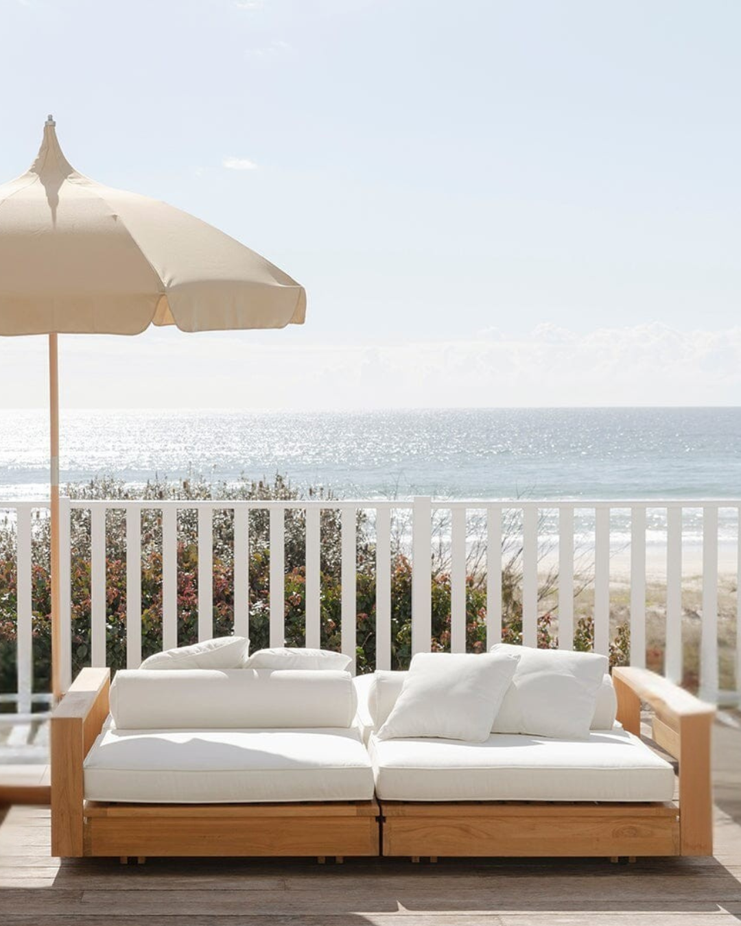 Teak outdoor double sunlounge with white cushions on a deck overlooking the ocean.