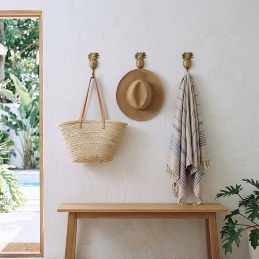 Three brass wall hooks hang a woven straw tote bag, a wide-brim straw hat, and a striped beach towel on a white stucco wall above a light wood console table. A window on the left shows greenery outside, and a plant is visible in the lower right corner.