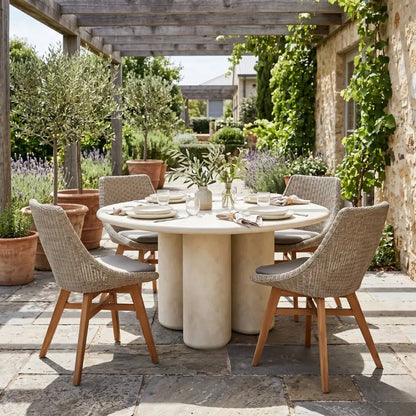 Outdoor dining setup with a round stone table and wicker chairs in a garden setting.