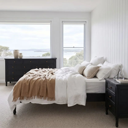 Neatly made bed with beige blanket and pillows in a bedroom with large windows.