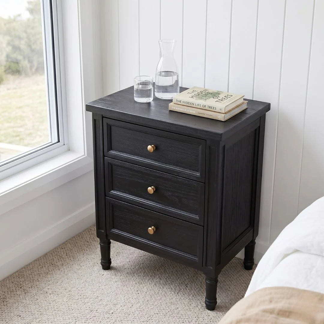 A black wooden three-drawer nightstand with gold knobs, styled with a glass carafe, water glass, and books on top, set in a bedroom corner next to a window.