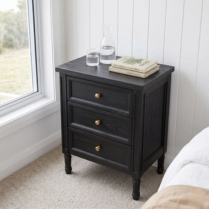 A black wooden three-drawer nightstand with gold knobs, styled with a glass carafe, water glass, and books on top, set in a bedroom corner next to a window.