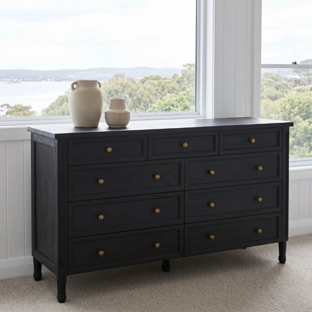 Black dresser with gold knobs in a room with large windows showing a view of trees and sky.