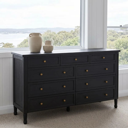 Black dresser with gold knobs in a room with large windows showing a view of trees and sky.
