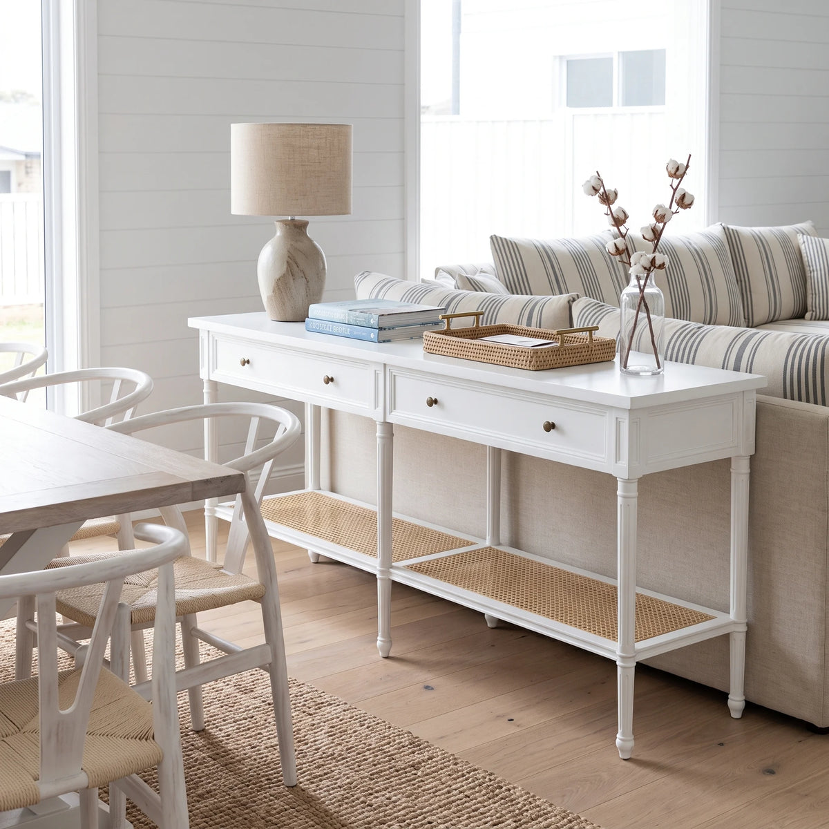 A white console table with two drawers, a lower woven cane shelf, and small brass knobs, styled in a bright coastal living room next to a sofa and dining table.