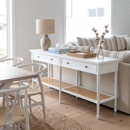 A white console table with two drawers, a lower woven cane shelf, and small brass knobs, styled in a bright coastal living room next to a sofa and dining table.