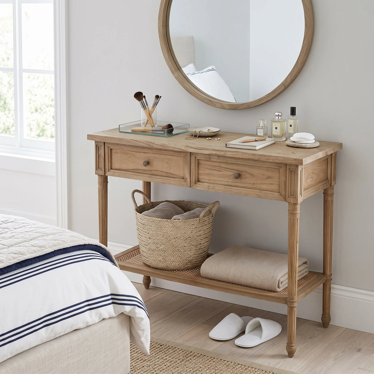 A light natural wood console table with two drawers, an open lower shelf, and a woven rattan seat, styled in a bedroom setting with a round mirror, toiletries, a wicker basket, folded towels, and white slippers.