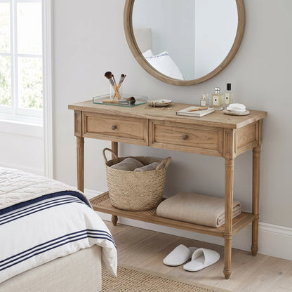 A light natural wood console table with two drawers, an open lower shelf, and a woven rattan seat, styled in a bedroom setting with a round mirror, toiletries, a wicker basket, folded towels, and white slippers.
