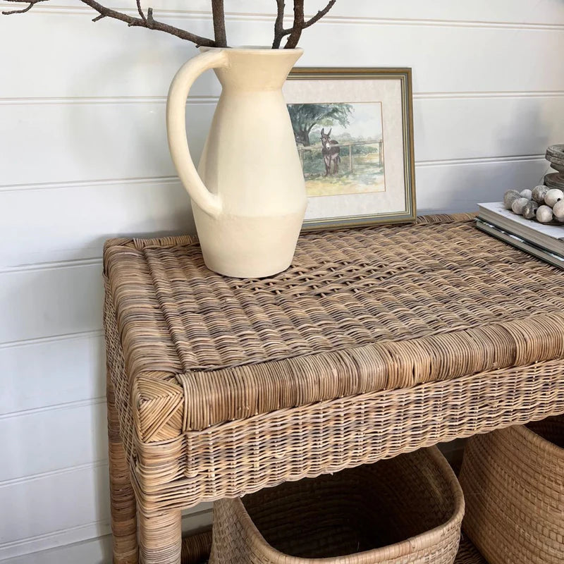 Wicker side table with a white pitcher on top, framed picture in the background