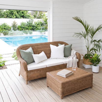 A wicker outdoor loveseat with white seat cushions and green and white pillows, paired with a matching wicker ottoman on a light deck beside a pool and potted plants.