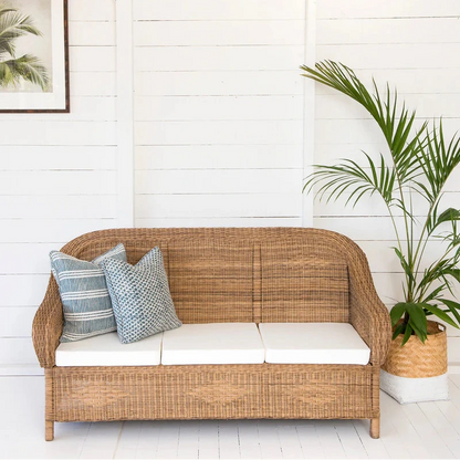 Wicker outdoor sofa with cushions against a white wooden wall with a plant.