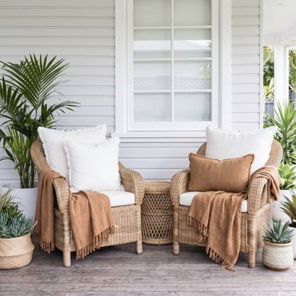Two woven rattan armchairs on a porch with white cushions, tan throw pillows, and draped tan fringed blankets, surrounded by potted plants.