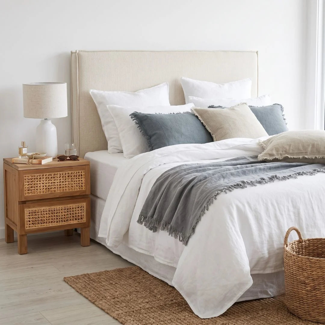 A styled bedroom scene showing a bed with white linens, gray and beige pillows, a gray throw blanket, a tall upholstered headboard, a wooden nightstand with a white lamp, and a woven basket on the floor.