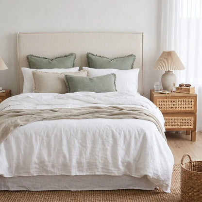 A neatly made bed with white linens, multiple beige and sage green pillows, a light beige upholstered headboard, a woven nightstand, and a matching lamp in a softly lit bedroom.