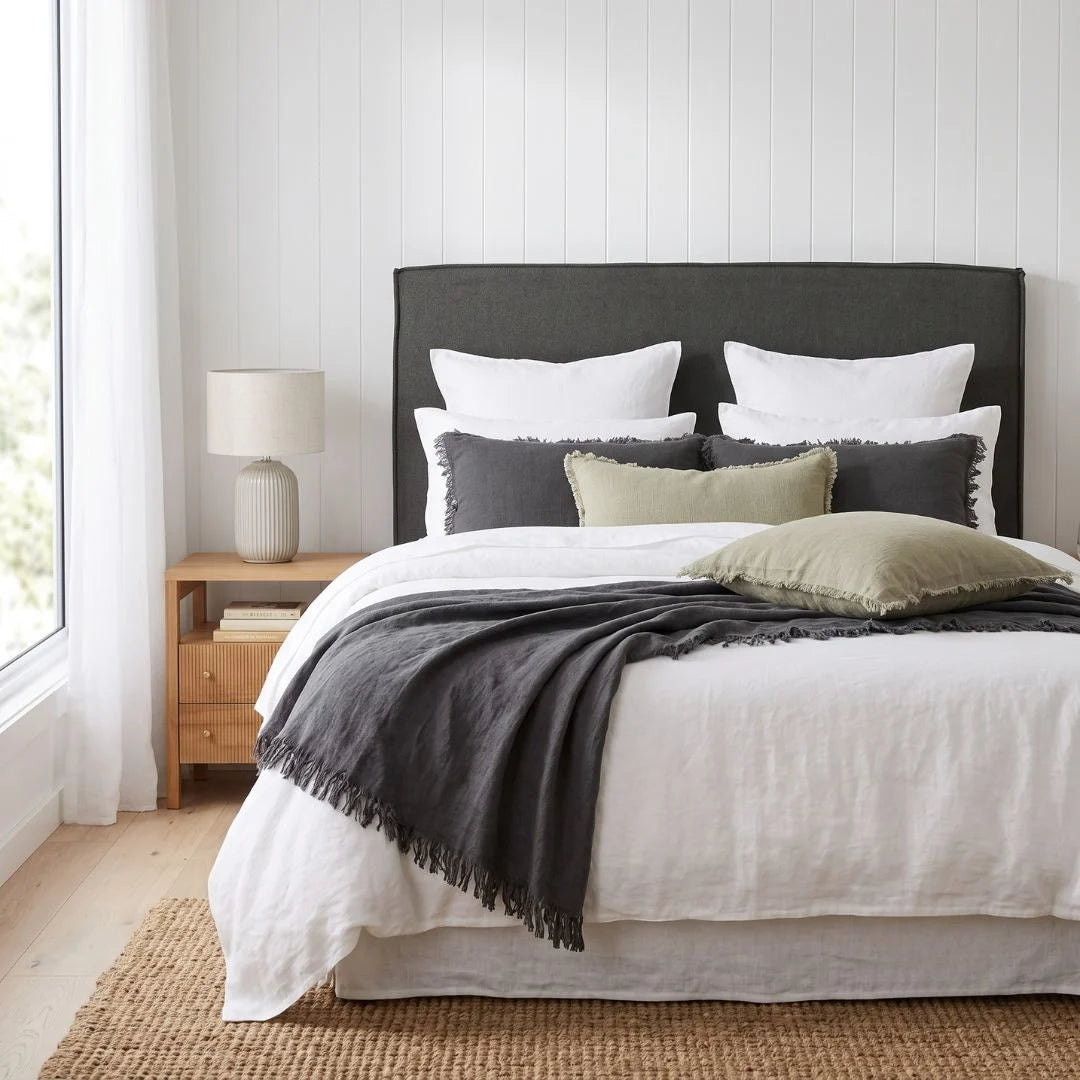 A neatly made bed in a bright bedroom setting, featuring a dark charcoal upholstered headboard, white bedding, gray and olive accent pillows, a dark gray throw blanket with fringe, a wooden nightstand with a table lamp, and a woven rug.