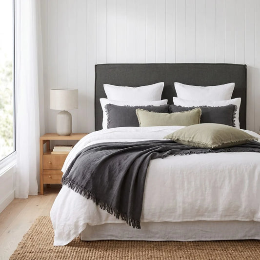 A neatly made bed in a bright bedroom setting, featuring a dark charcoal upholstered headboard, white bedding, gray and olive accent pillows, a dark gray throw blanket with fringe, a wooden nightstand with a table lamp, and a woven rug.