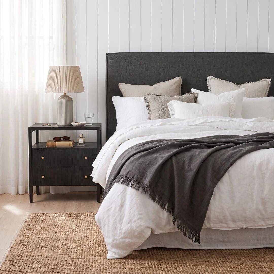 A styled bedroom scene featuring a bed with white linens, a dark charcoal gray upholstered headboard, matching gray throw blanket, beige and white pillows, a black nightstand with a lamp, and a woven jute rug.