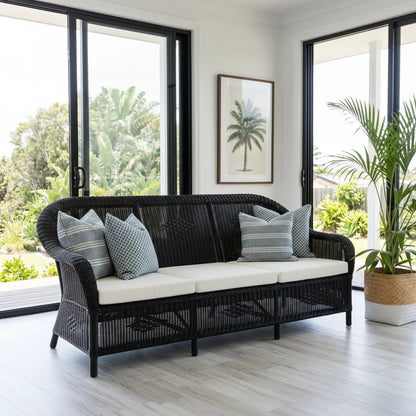 Black wicker sofa with white cushions in a room with large windows and plants.