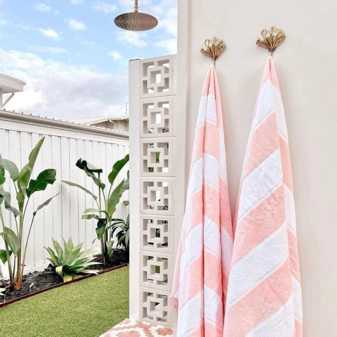 Two pink and white striped towels hanging on gold hooks against a white wall with outdoor plants and a shower head in the background.