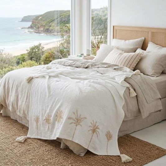 Neatly made bed with beige bedding featuring palm tree designs, in a room with large windows overlooking a scenic view.
