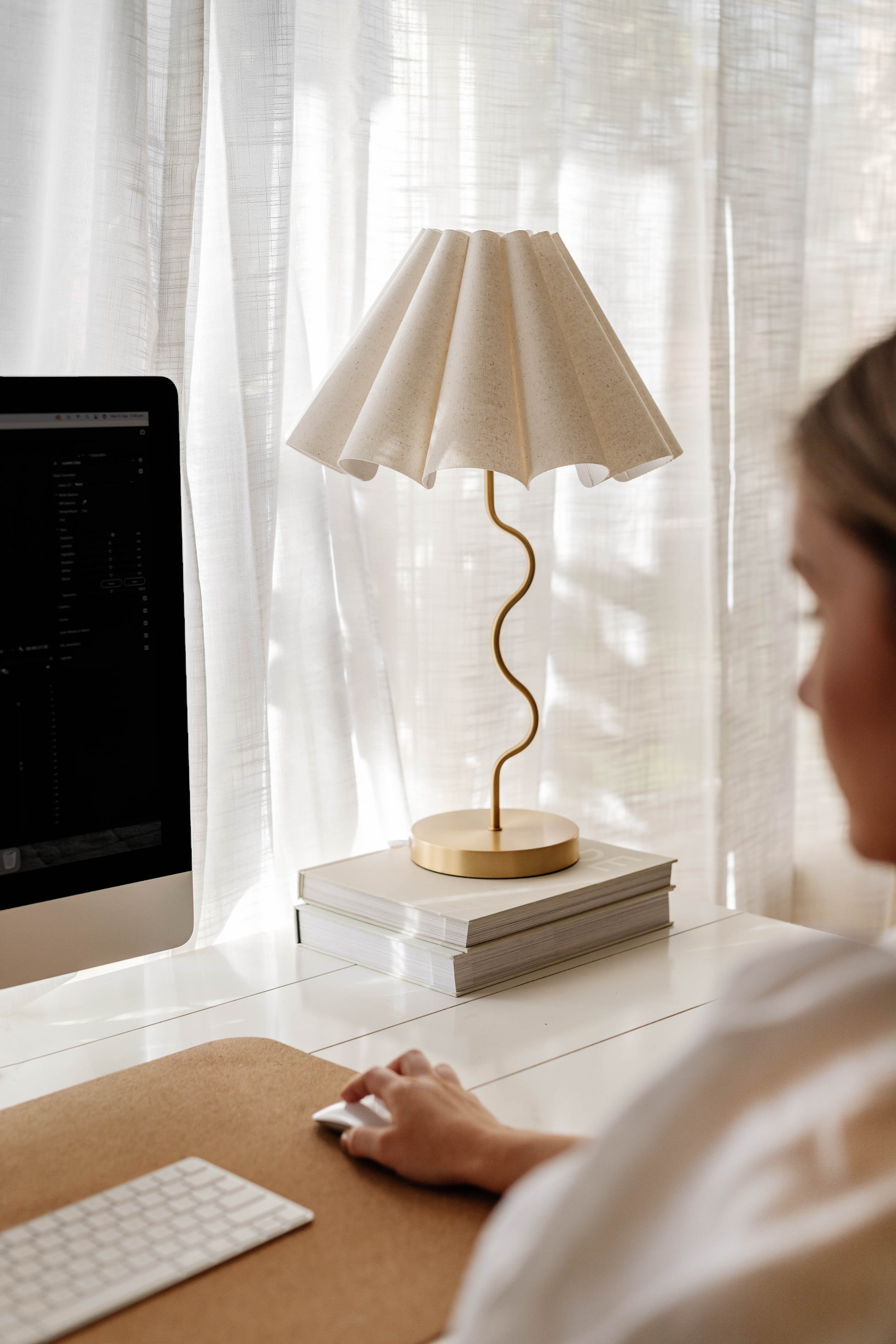 Person using a computer at a desk with a lamp and books in the background