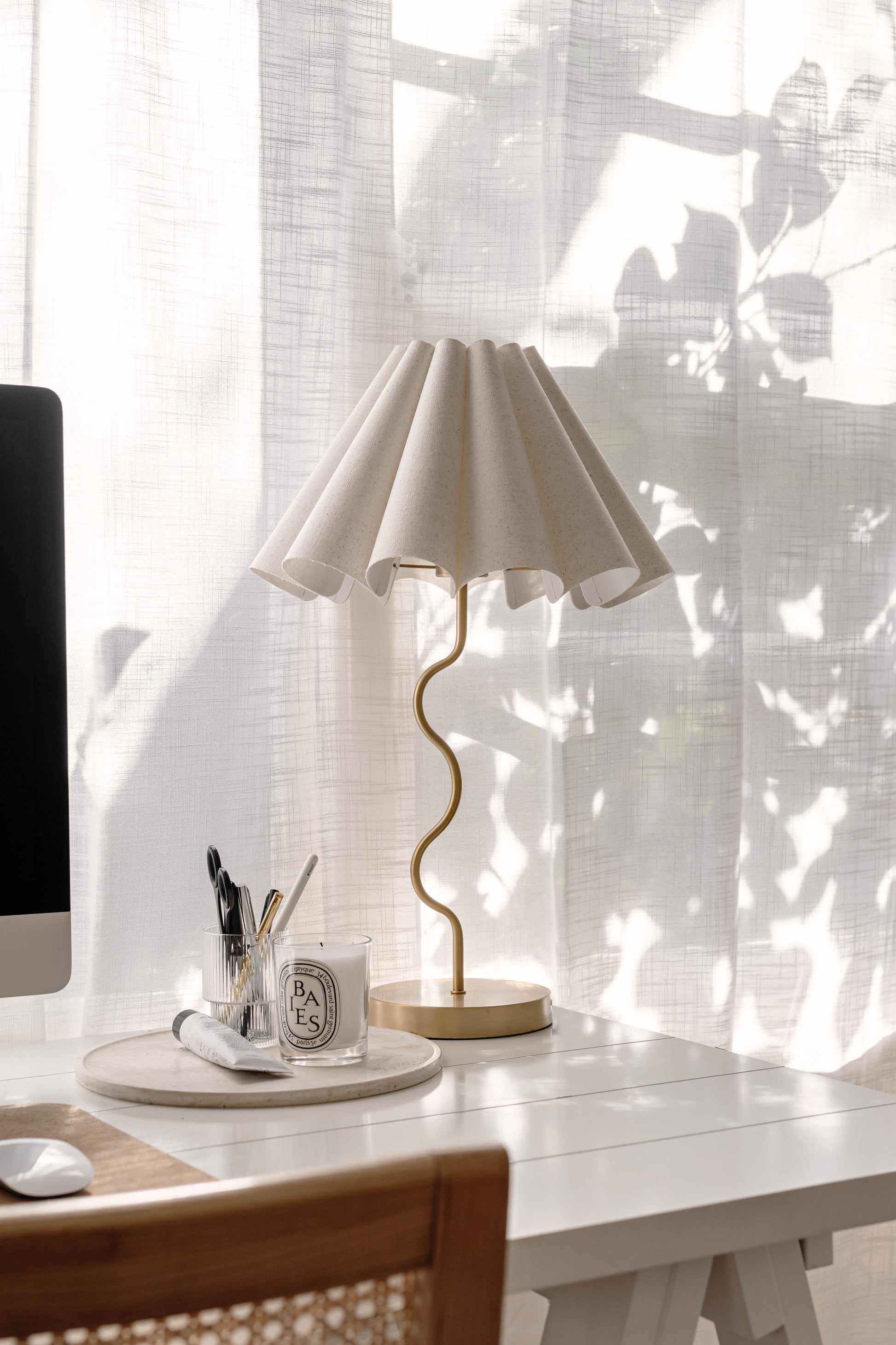 Desk with a lamp, mug, and computer in a bright room with sheer curtains.