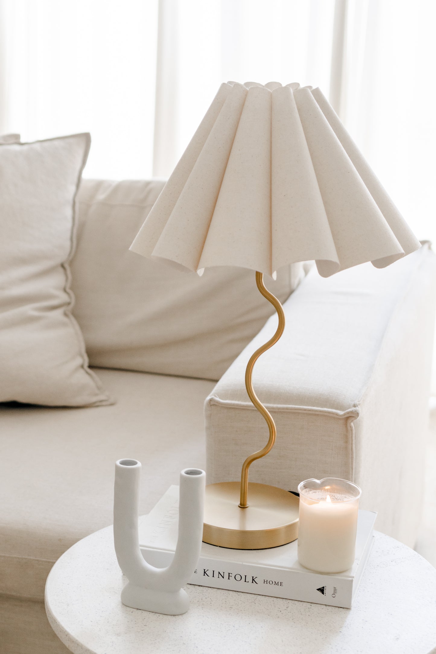 White lamp with gold base on a table next to a candle and decorative book in a living room setting.