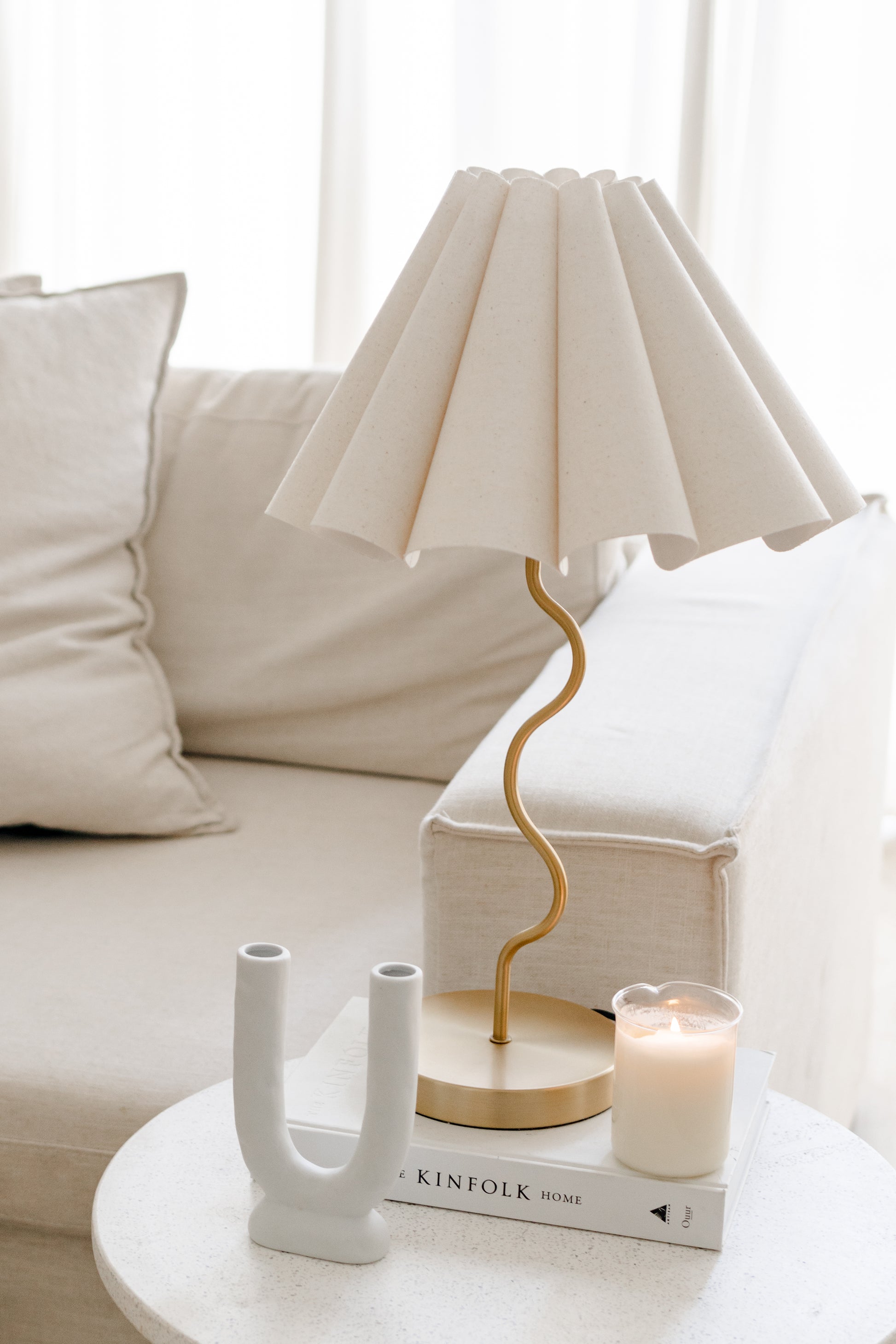 White lamp with gold base on a table next to a candle and decorative book in a living room setting.