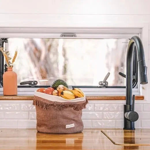 Caravan kitchen sink area with a window above, showing a view of trees, a basket of fruits and vegetables on the windowsill, and a black faucet.